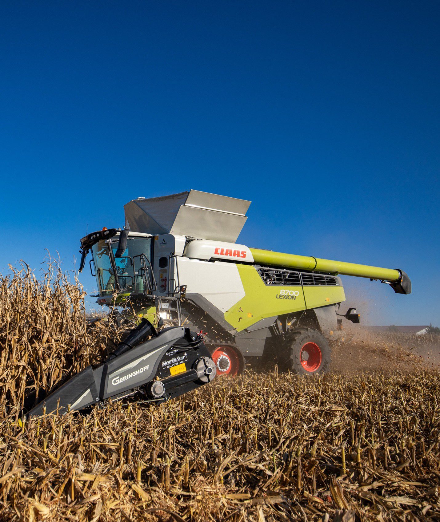 Large green-and-white CLAAS 8700 Lexion combine harvester with front Geringhoff corn header working through a dry cornfield under a clear blue sky, illustrating precision farming automation and connected agricultural equipment to boost productivity and reduce costs